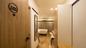 Quarto de hotel com porta de madeira, cama com colcha branca, escrivaninha com cadeira e unidade de ar condicionado no teto.