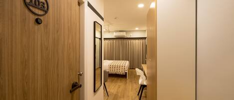 A hotel room with a wooden door, a bed with a white bedspread, a desk with a chair, and an air conditioning unit on the ceiling.