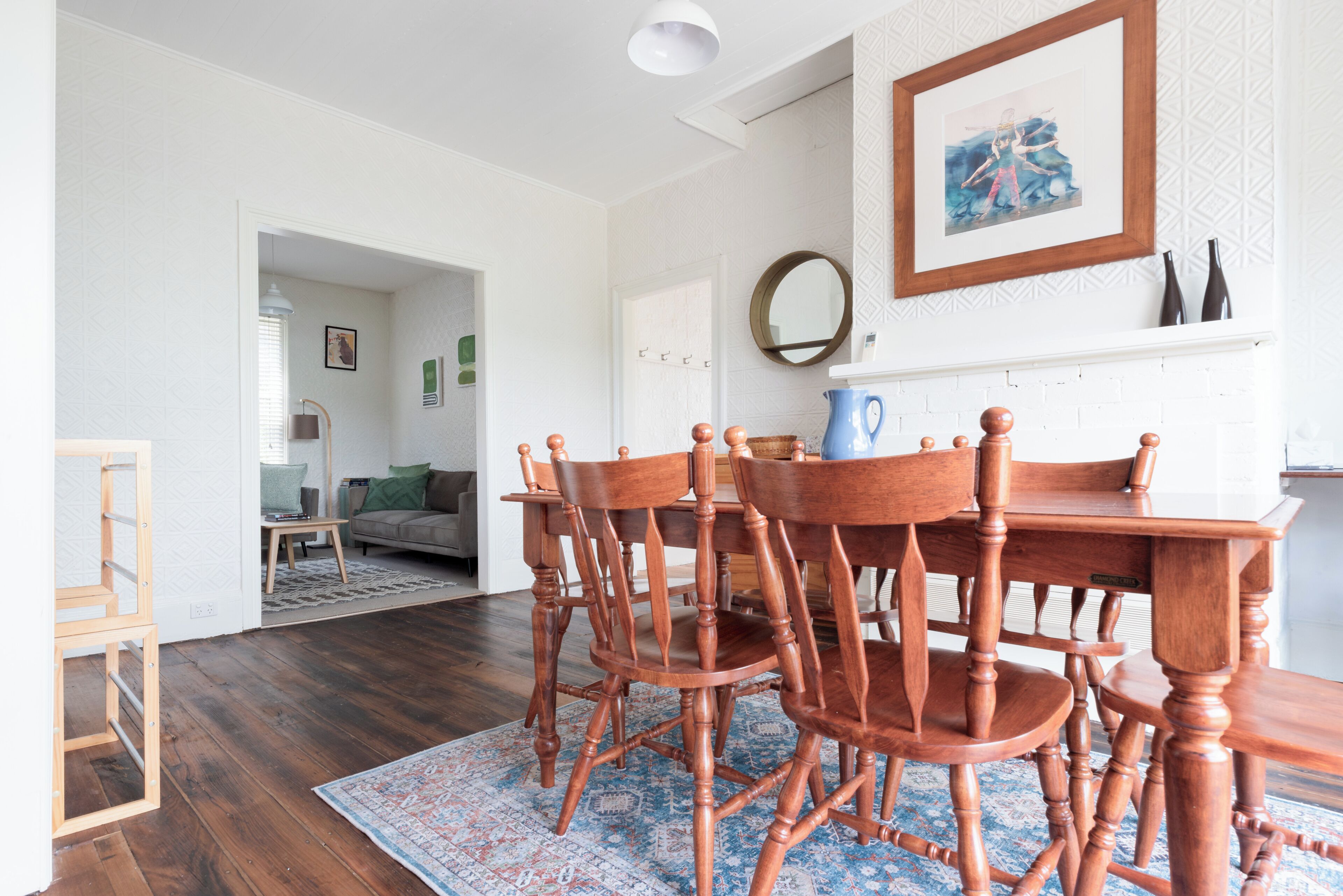 A dining area with a wooden table and chairs, a blue patterned rug, and a white wall with a framed picture.