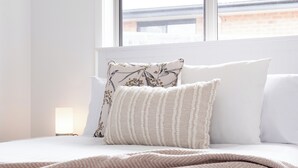 A neatly made bed with a patterned throw blanket and two pillows, one with a floral design and the other striped, in a room with a large window. - The Cosy Spot - with Parking (Mowbray)