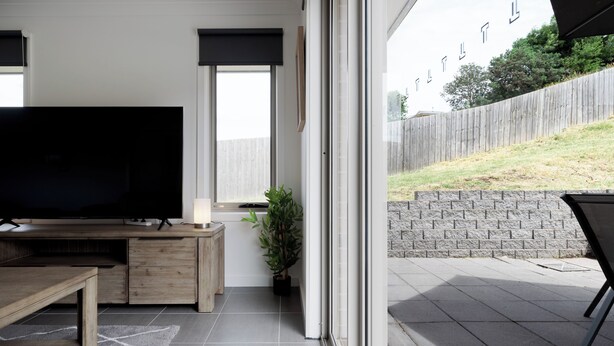 A modern living room with a large flat-screen TV, wooden furniture, a potted plant, and a sliding glass door leading to a patio with a stone wall and a wooden fence. - The Cosy Spot - with Parking (Mowbray)