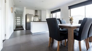 A modern kitchen with a central island, a dining area with a wooden table and dark chairs, and a view of the outdoors through large windows. - The Cosy Spot - with Parking (Mowbray)