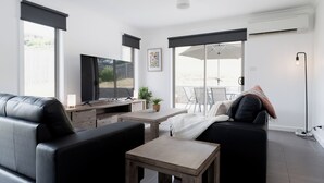 A modern living room with a black leather sofa, a wooden coffee table, and a TV on a wooden stand. There is a small table with a plant, and a large window with a view of the outdoors. - The Cosy Spot - with Parking (Mowbray)