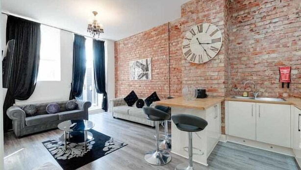 A modern living room with a brick wall, a large clock, a sofa, a coffee table, and a kitchen area with a sink and cabinets. - Dukes Street Apartment Hotel (Liverpool)