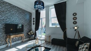 A modern living room with a stone-patterned wall, a large flat-screen TV, a glass coffee table, a black sofa, and a chandelier. - Dukes Street Apartment Hotel (Liverpool)