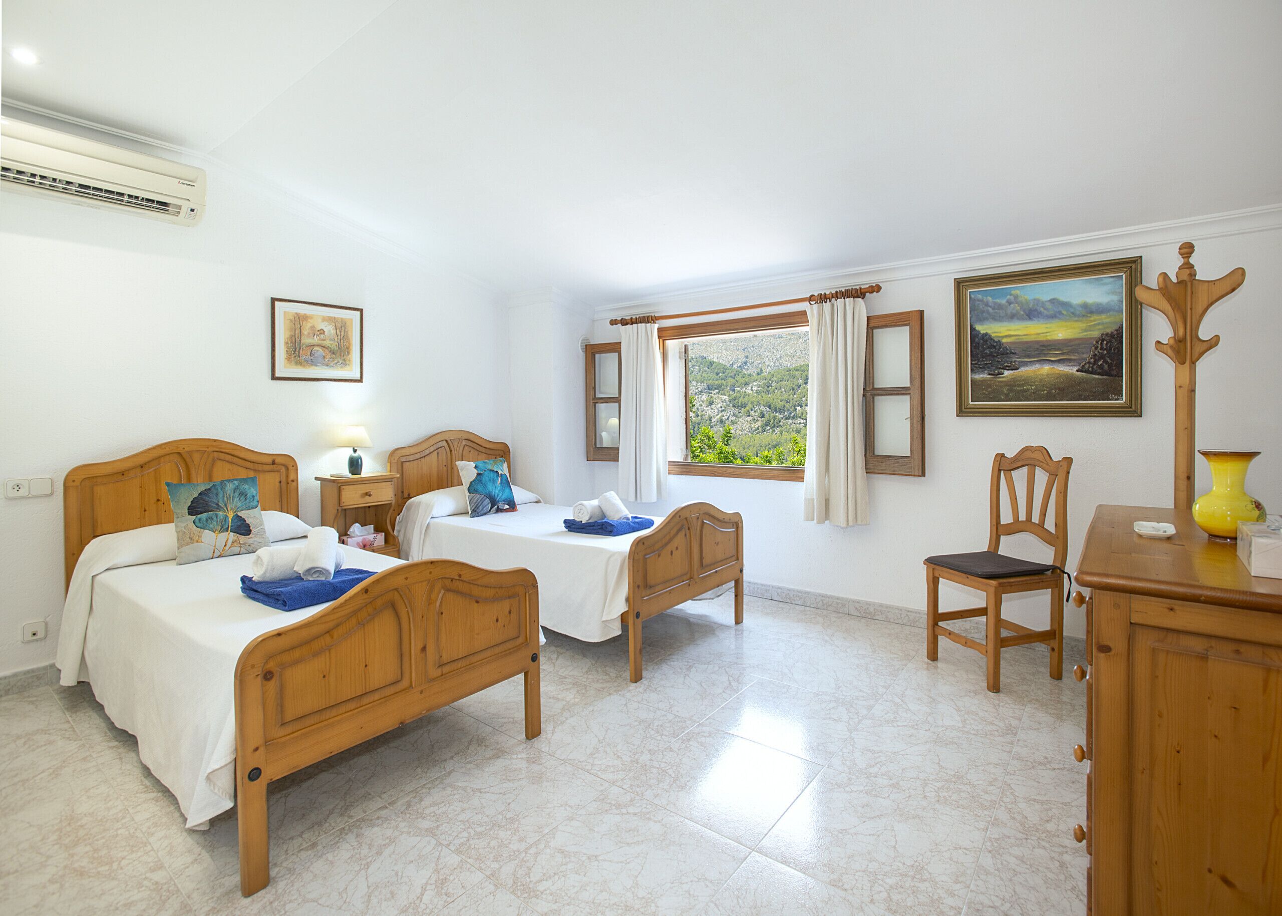 Habitación de hotel con dos camas de madera, una silla de madera, un escritorio, una ventana con cortinas y un cuadro en la pared.