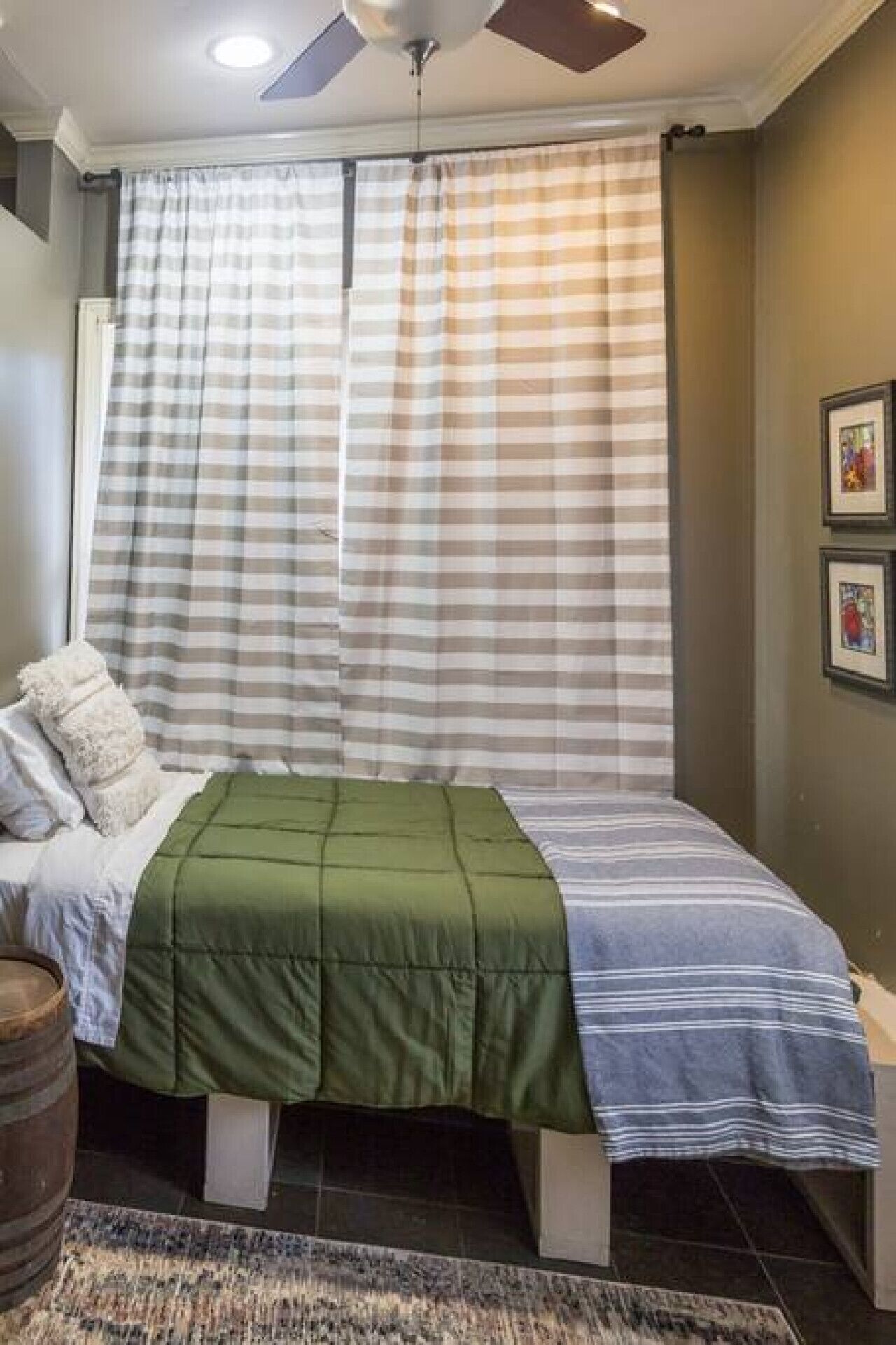 A bedroom with a bed, a green and blue striped bedspread, a wooden barrel, and a patterned rug.