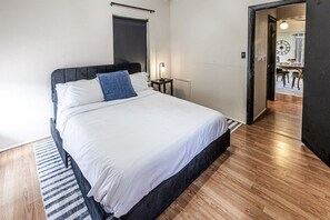 A neatly made bed with white linens and a blue accent pillow in a room with wooden flooring and a striped rug. - Trailside Retreat in the City (Tulsa)