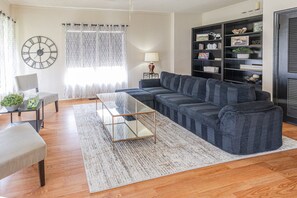 Ein Wohnzimmer mit einem Ecksofa, einem Glastisch, einem Beistelltisch, einem Sessel, einem Bücherregal und einer Wanduhr.