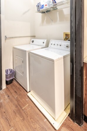 A laundry room with two front-loading washing machines, a shelf with cleaning supplies, and a trash bin. - Trailside Retreat in the City (Tulsa)