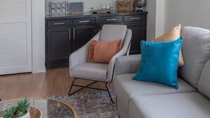 Una sala de estar con un sofá gris, una mesa de centro redonda de vidrio, un tapete estampado, un mueble de madera oscura con estantes, y una puerta blanca.