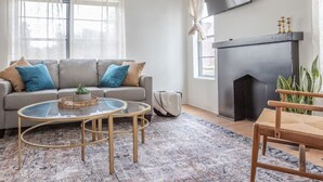 Una sala de estar con un sofá gris, una mesa de centro de vidrio, una chimenea y un televisor montado en la pared.