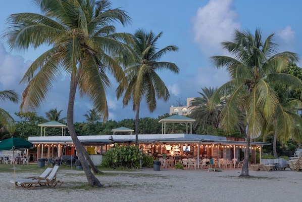 A beachfront restaurant with outdoor seating, palm trees, and a covered area.