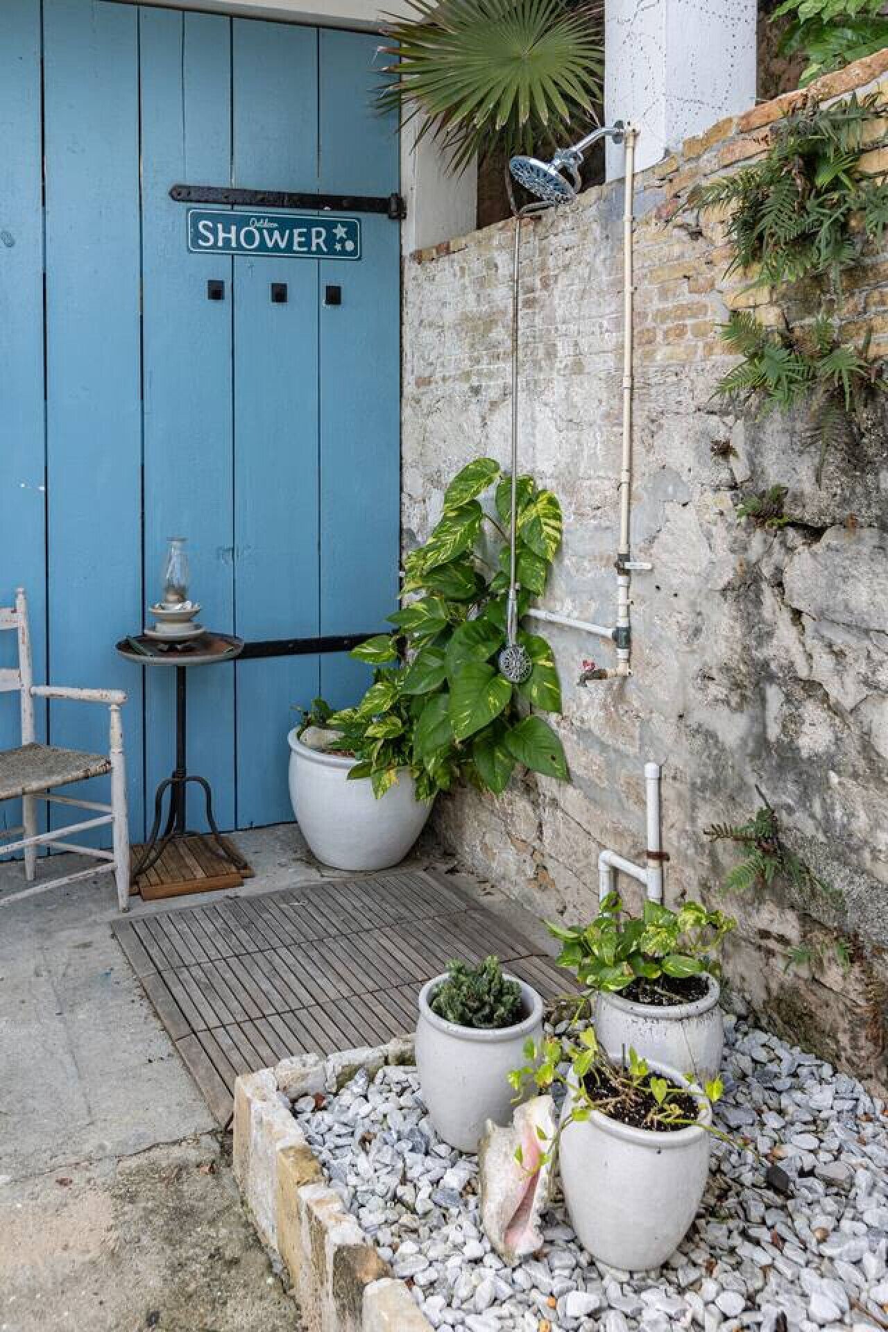 Une porte bleue portant l’inscription « DOUCHE », une chaise blanche et des plantes en pot dans une cour.