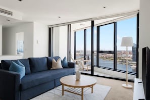 A modern living room with a blue sofa, a round wooden coffee table, and a large window offering a city view.