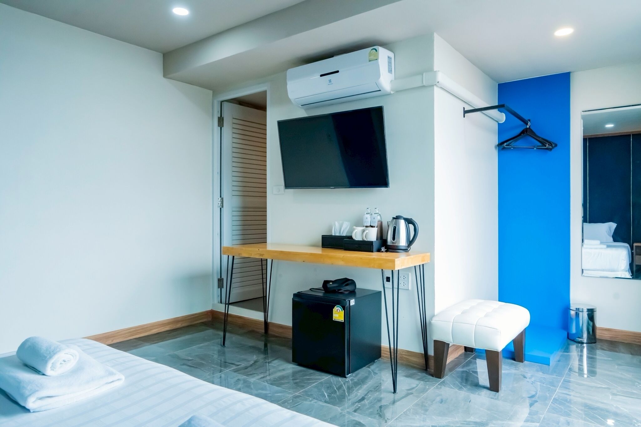 A modern hotel room with a flat-screen TV, a small table with a kettle, a mini fridge, a white cushioned bench, and a blue accent wall.