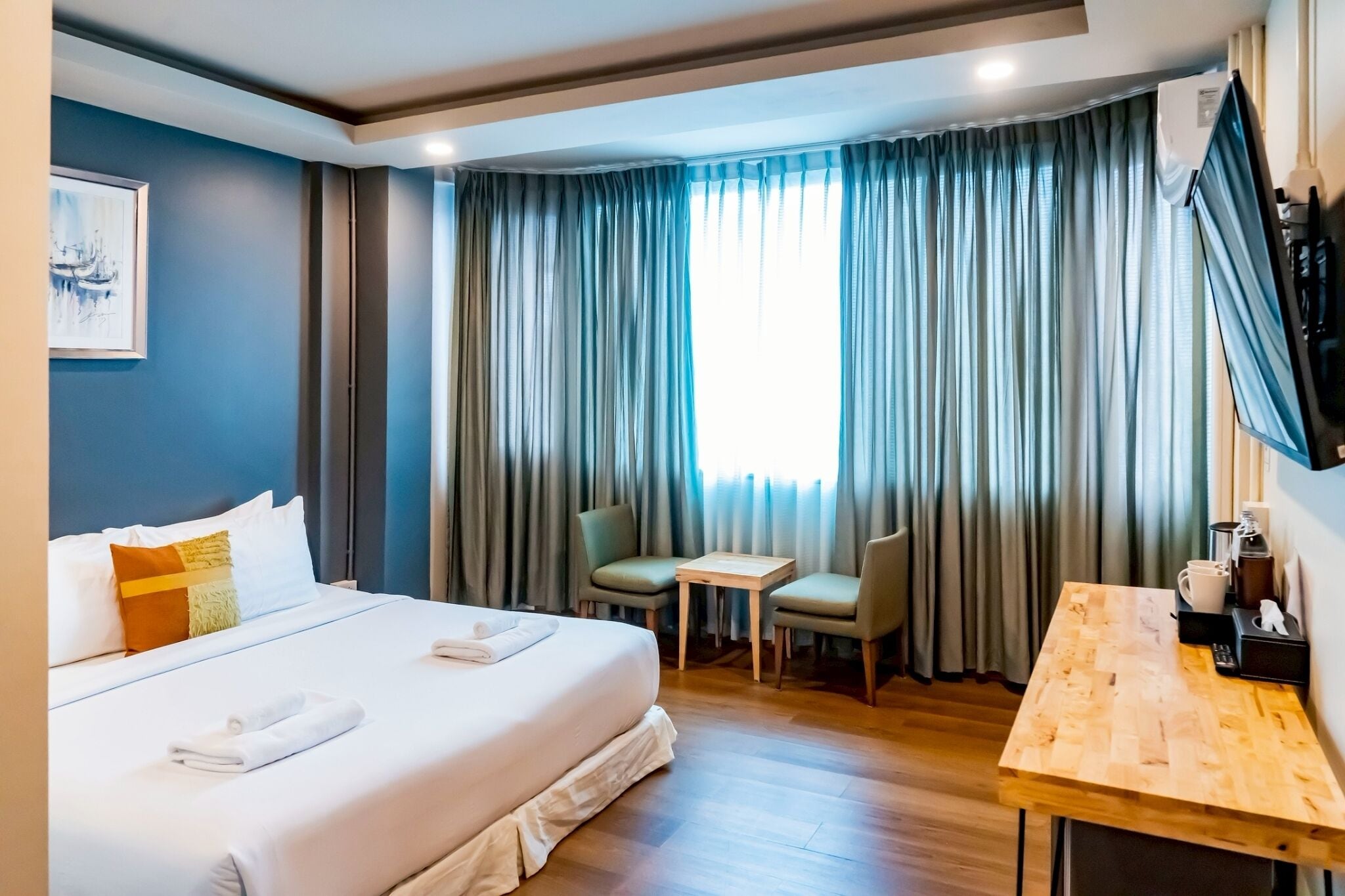 A hotel room with a bed, a small table, two chairs, a TV mounted on the wall, and a window with curtains.