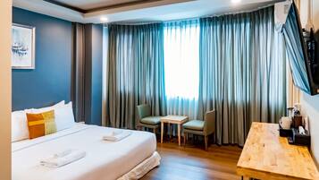 A hotel room with a bed, a small table, two chairs, a TV mounted on the wall, and a window with curtains.