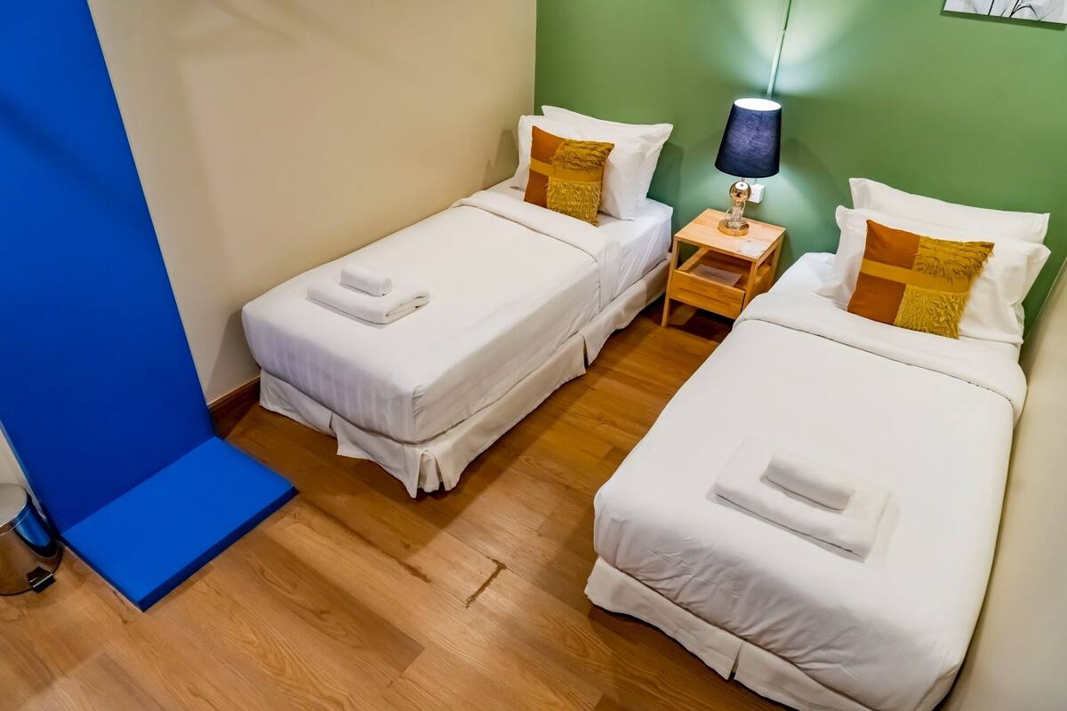Una habitación de hotel con dos camas individuales, una mesita de noche pequeña con lámpara y una silla azul.