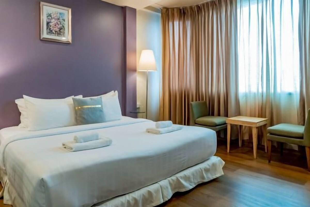 A hotel room with a bed, a desk, two chairs, a lamp, and a window with curtains.