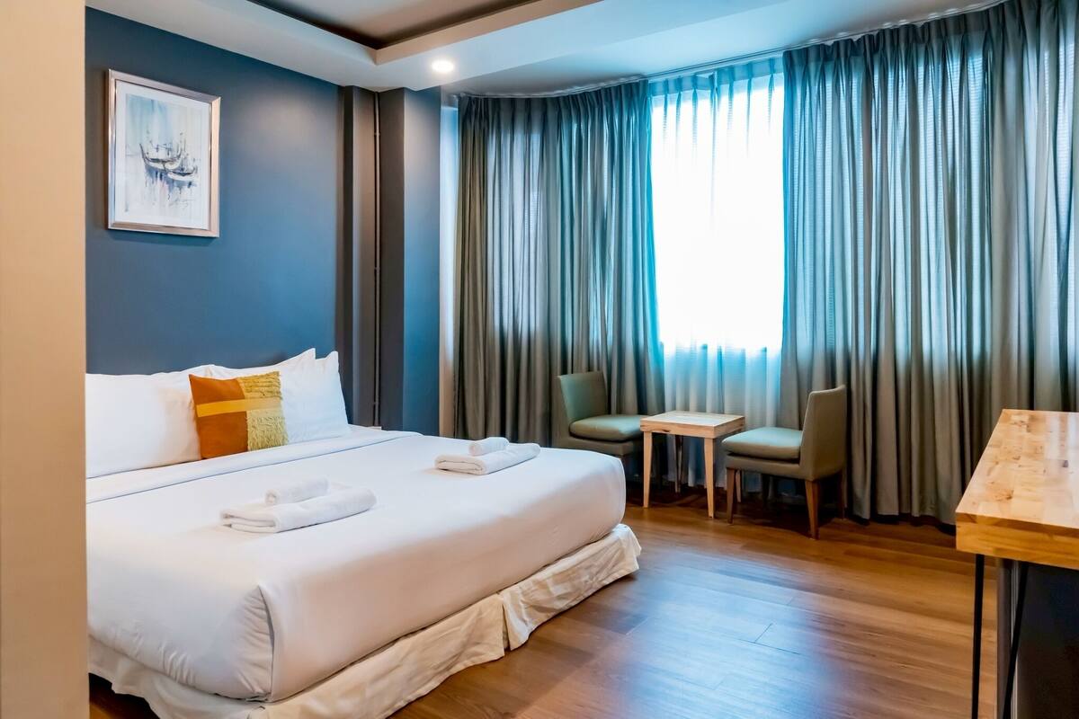 Habitación de hotel con una cama grande, una mesita pequeña, una silla y una ventana con cortinas.