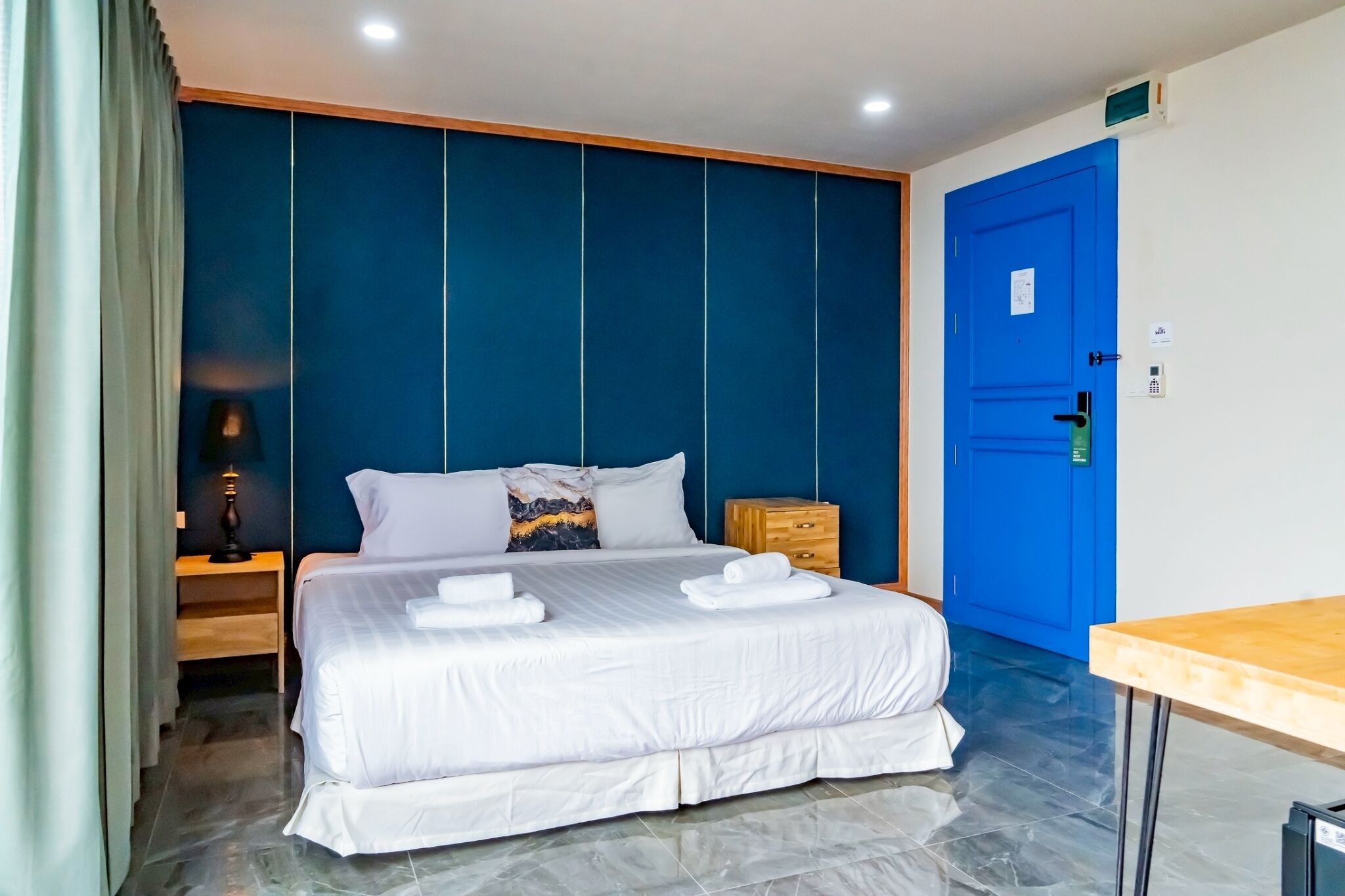 Habitación de hotel con una cama grande, una mesita de noche de madera, una puerta azul y una pequeña mesa.