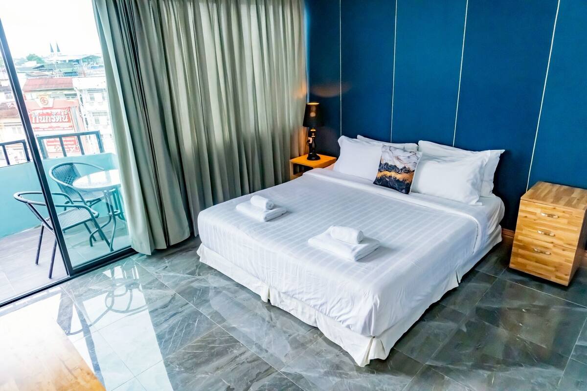 Habitación de hotel con una cama grande, una mesita de noche de madera, una mesa de vidrio con sillas y vistas a edificios a través de la ventana.