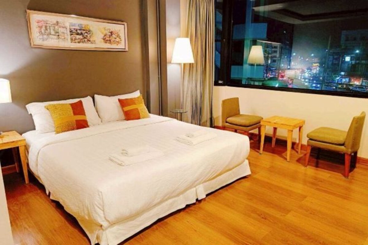 Habitación de hotel con una cama grande, un escritorio, dos sillas, una lámpara y una ventana con vistas a la ciudad.