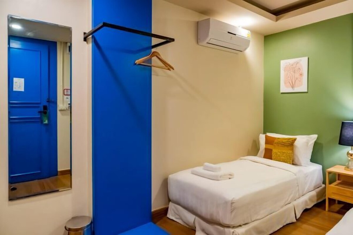 A hotel room with a bed, a hanging clothes rack, a small bedside table, and a wall-mounted air conditioning unit.