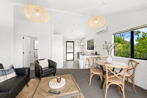 Uma sala de estar moderna com um sofá de couro preto, uma mesa de centro de madeira com um controle remoto, e uma área de jantar com mesa e cadeiras de madeira.