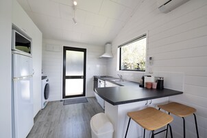 Cozinha compacta com máquina de lavar, fogão e pia. Há duas banquetas de madeira e uma janela que ilumina naturalmente.