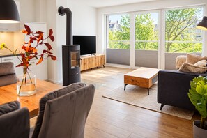 A modern living room with a wood-burning stove, a dining area with a vase of flowers, and large windows offering a view of the outdoors. - Vacation rental (new construction 2023) in prime location with fireplace and bal (Winterberg)