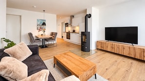 A modern living room with a sofa, coffee table, and a TV. There's a dining area with a table and chairs. A kitchen with cabinets and a stove is visible. - Vacation rental (new construction 2023) in prime location with fireplace and bal (Winterberg)