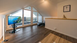 Un salon moderne doté d’un grand téléviseur, d’un plancher en bois, offrant une vue dégagée grâce à de grandes fenêtres, et orné d’un tableau encadré au mur.