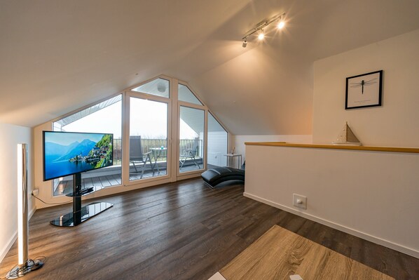 Ein modernes Wohnzimmer mit großem Fernseher, Holzboden, Blick durch hohe Fenster und einem gerahmten Bild an der Wand.