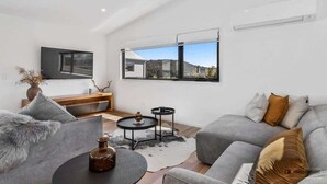 Un salon moderne avec un canapé gris, une table basse, un téléviseur fixé au mur et une grande fenêtre donnant sur un immeuble et des collines.