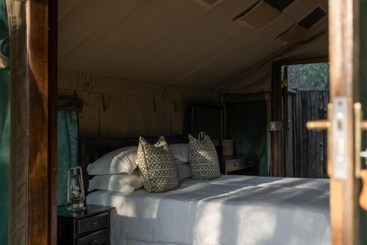 A bed with patterned pillows, a bedside table with a lamp, and a curtain partially covering the window.