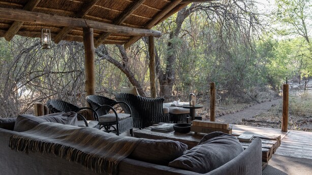 A covered outdoor seating area with wicker furniture and a wooden table, surrounded by trees. - Mashatu Tent Camp - Mashatu Game Reserve (Lentswelemoriti)