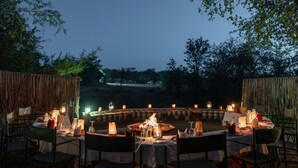 An outdoor dining area with a circular fire pit surrounded by tables and chairs, set against a backdrop of trees and a distant view of animals. - Mashatu Tent Camp - Mashatu Game Reserve (Lentswelemoriti)