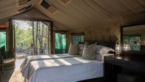 A spacious bedroom with a large bed, a wooden nightstand, a mirror, and a view of the outdoors. - Mashatu Tent Camp - Mashatu Game Reserve (Lentswelemoriti)