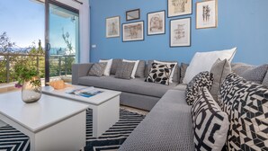 Ein modernes Wohnzimmer mit einem grauen Sofa, einem weißen Couchtisch und einem großen Fenster mit Blick ins Freie.