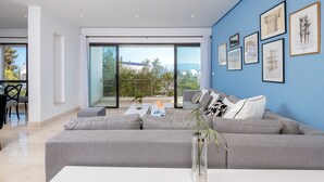 Ein modernes Wohnzimmer mit einem großen, grau-weißen Sofas, einer Glastür, die auf einen Balkon führt, und gerahmten Kunstwerken an der blauen Wand.