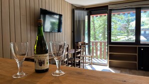 A wooden table with a bottle of wine and two empty glasses. A television is mounted on the wall, and a balcony with a view of trees is visible. - Bright studio in Pra-Loup with private parking, close to slopes (Pra-Loup)