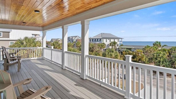 A covered porch with wooden flooring and white railings, overlooking a residential area with houses and a beach.