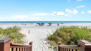 A sandy beach with a clear sky, a pathway leading to the beach, and several blue umbrellas.
