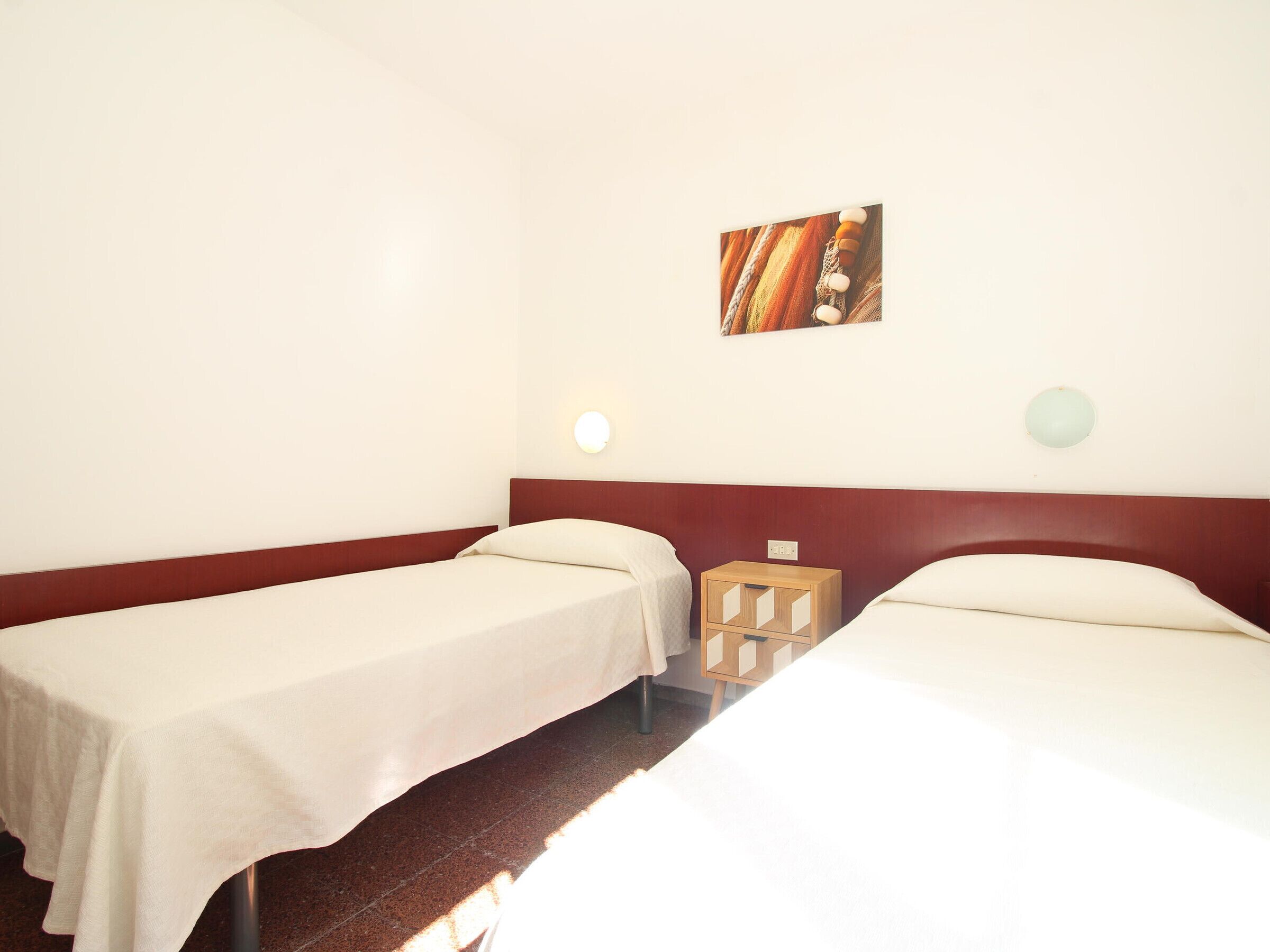 Une chambre d’hôtel avec deux lits simples, une table de chevet en bois, une lampe fixée au mur et un tableau.