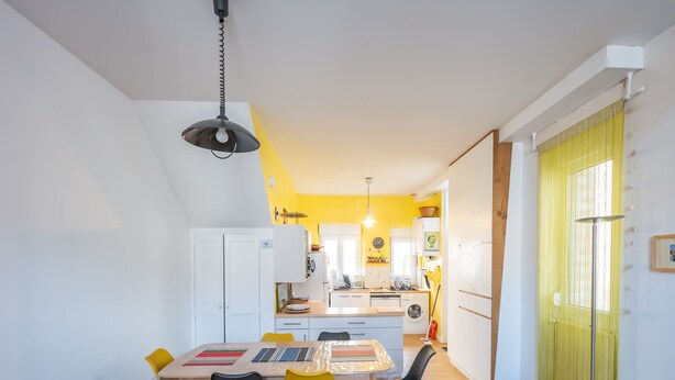 Una cocina compacta con armarios blancos, un comedor con mesa y sillas, y una pared amarilla.