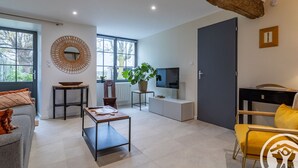 Una sala de estar moderna con un sofá, una mesa de centro y un televisor sobre un mueble. Hay un ventanal grande, una planta y una puerta en la habitación.