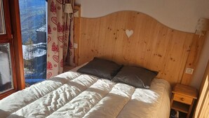 A bedroom with a wooden headboard, a bed with white bedding, two pillows, a nightstand, and a window with a view of a snowy landscape.