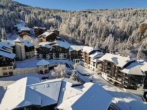 Une station de ski enneigée, composée de plusieurs bâtiments, dont une tour, et s’étendant au pied d’une montagne boisée.